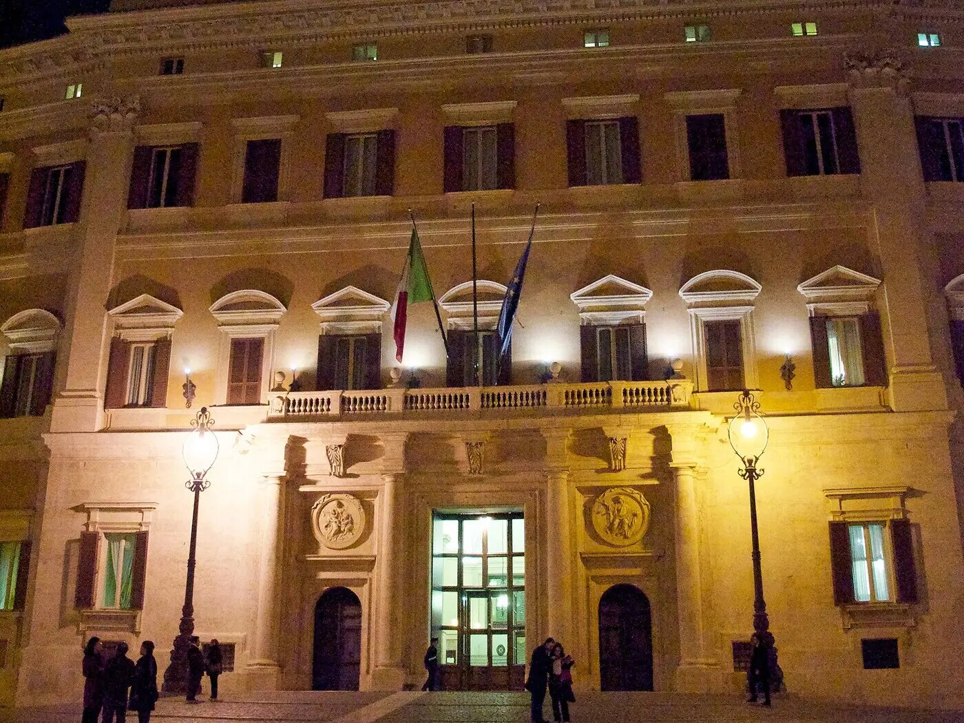 罗马众议院所在地蒙特奇托里奥宫(Palazzo Montecitorio)夜景,阳台上挂着意大利国旗和欧盟旗,门前两盏路灯、入口亮灯,广场上有几名行人;资料图,摄于2010年3月19日(图:Goldmund100 / CC BY-SA 3.0),已裁切